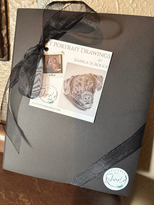 Black folder with black ribbon and portrait drawings by Sharla Scroggs on a textured surface.